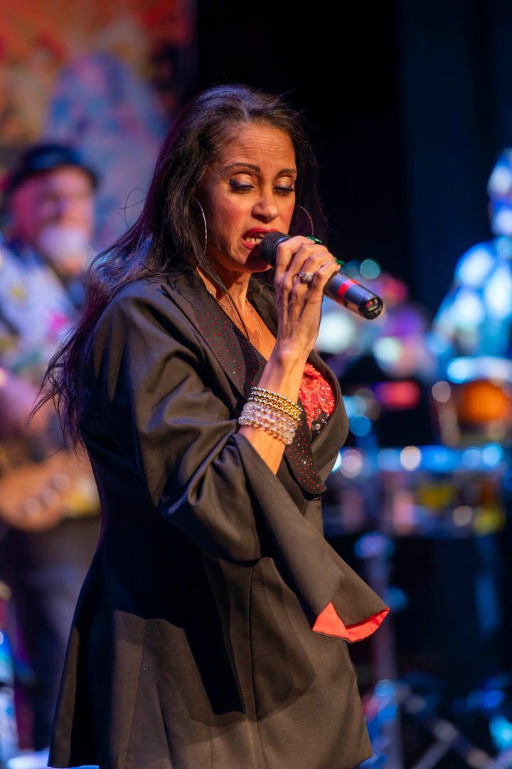 Singer performing onstage with a microphone, wearing a black outfit under colorful stage lights.