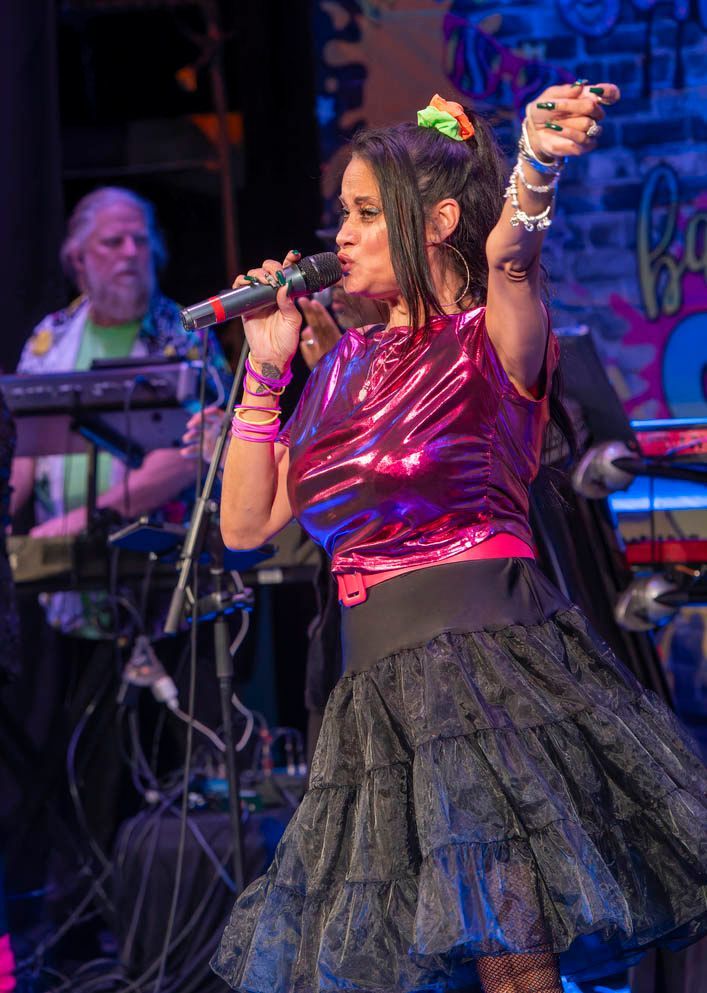 Singer in a shiny pink top and black skirt performing onstage with microphone, arm raised.