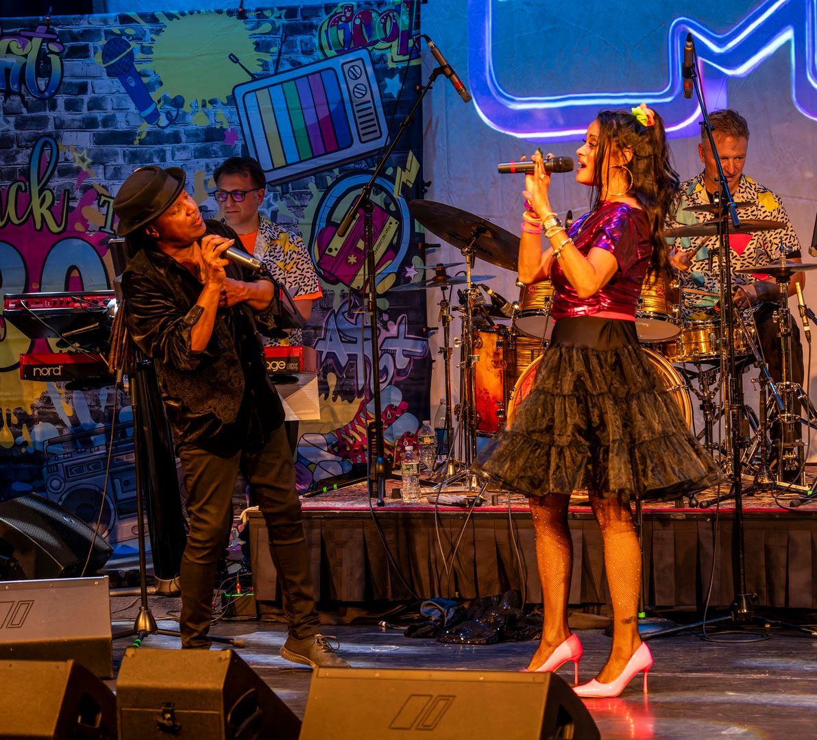 Two singers performing onstage with a band and neon graffiti backdrop