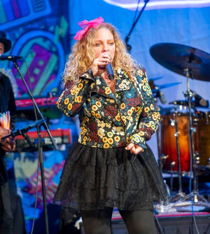 Singer performing onstage with a microphone, wearing a floral jacket and black tulle skirt, under colorful lights