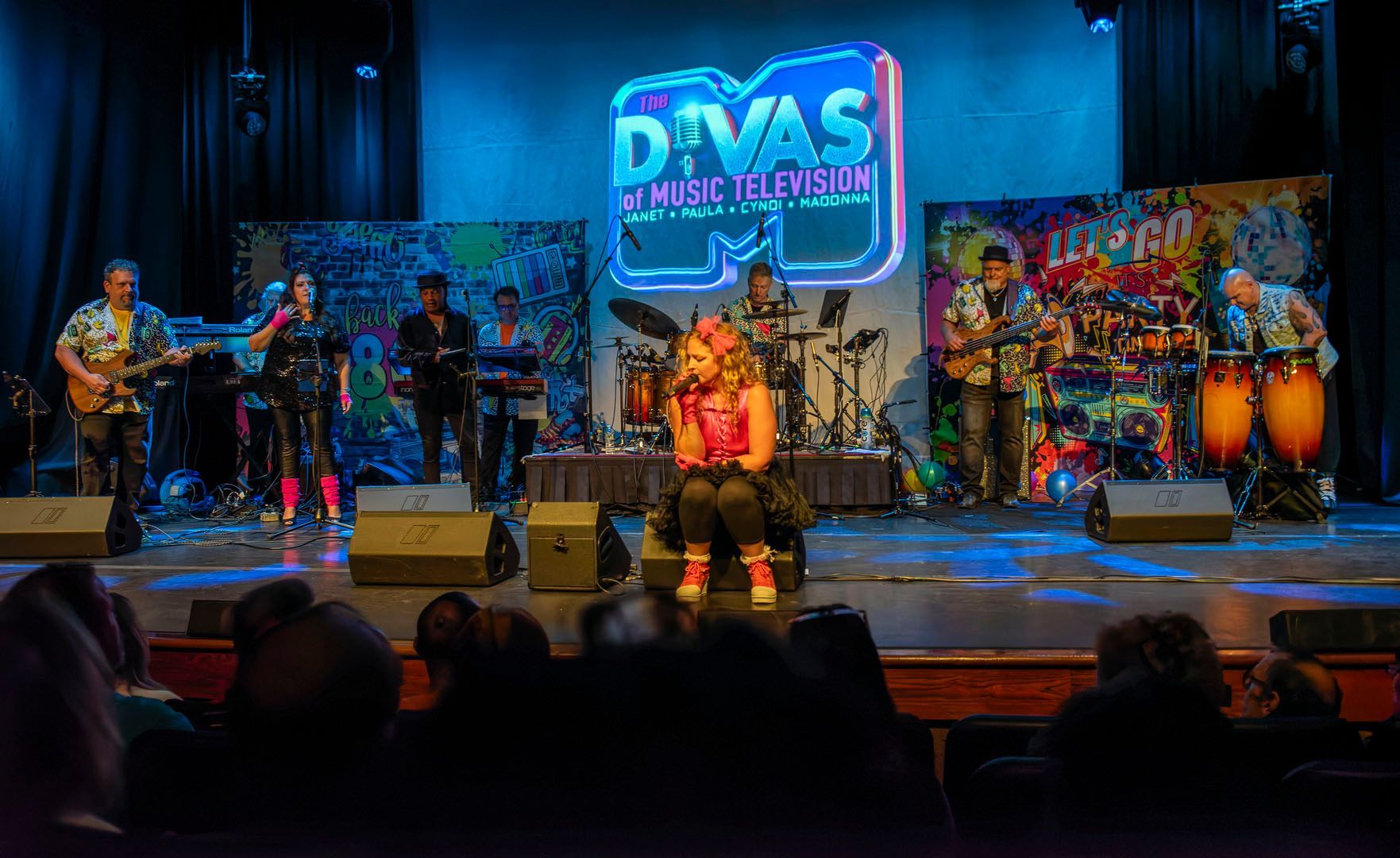 Live stage band performance with colorful backdrop, singers, instruments, and audience silhouettes.