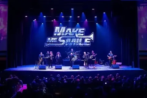 Blue-lit concert stage with performers beneath a large “MAKE” sign and seated audience in front