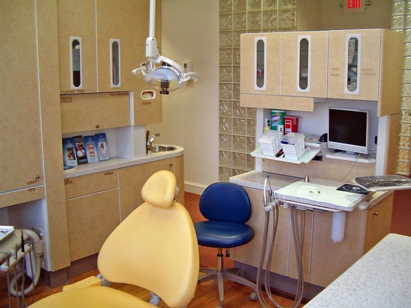 Empty dental exam room with chair, overhead lamp, cabinets, and computer workstation.