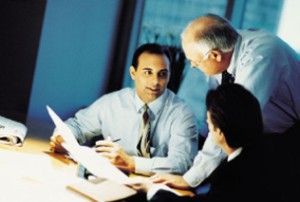 Three people in an office meeting, discussing documents at a table.