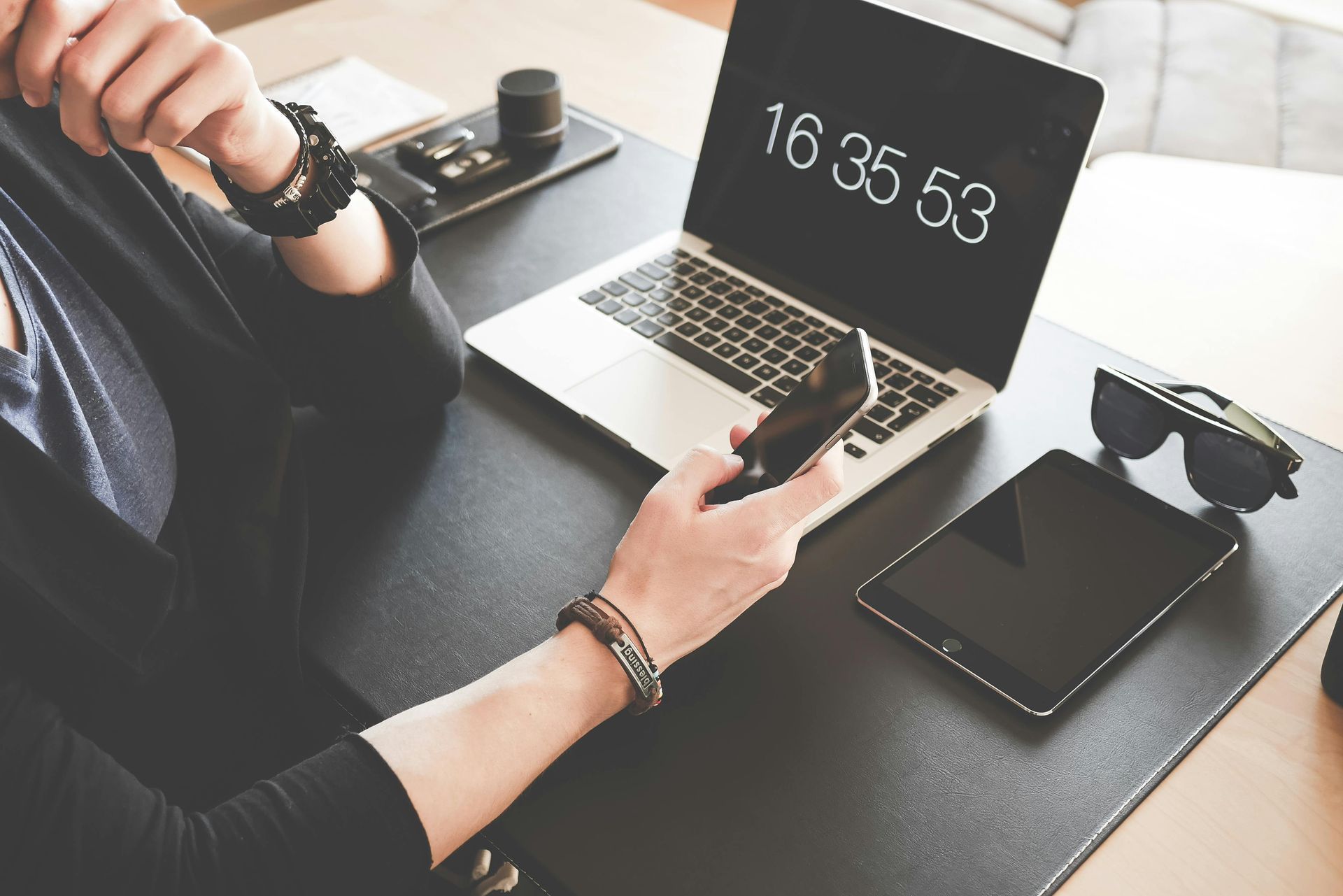 Person at desk with laptop, phone, and tablet. Time displayed on laptop screen.