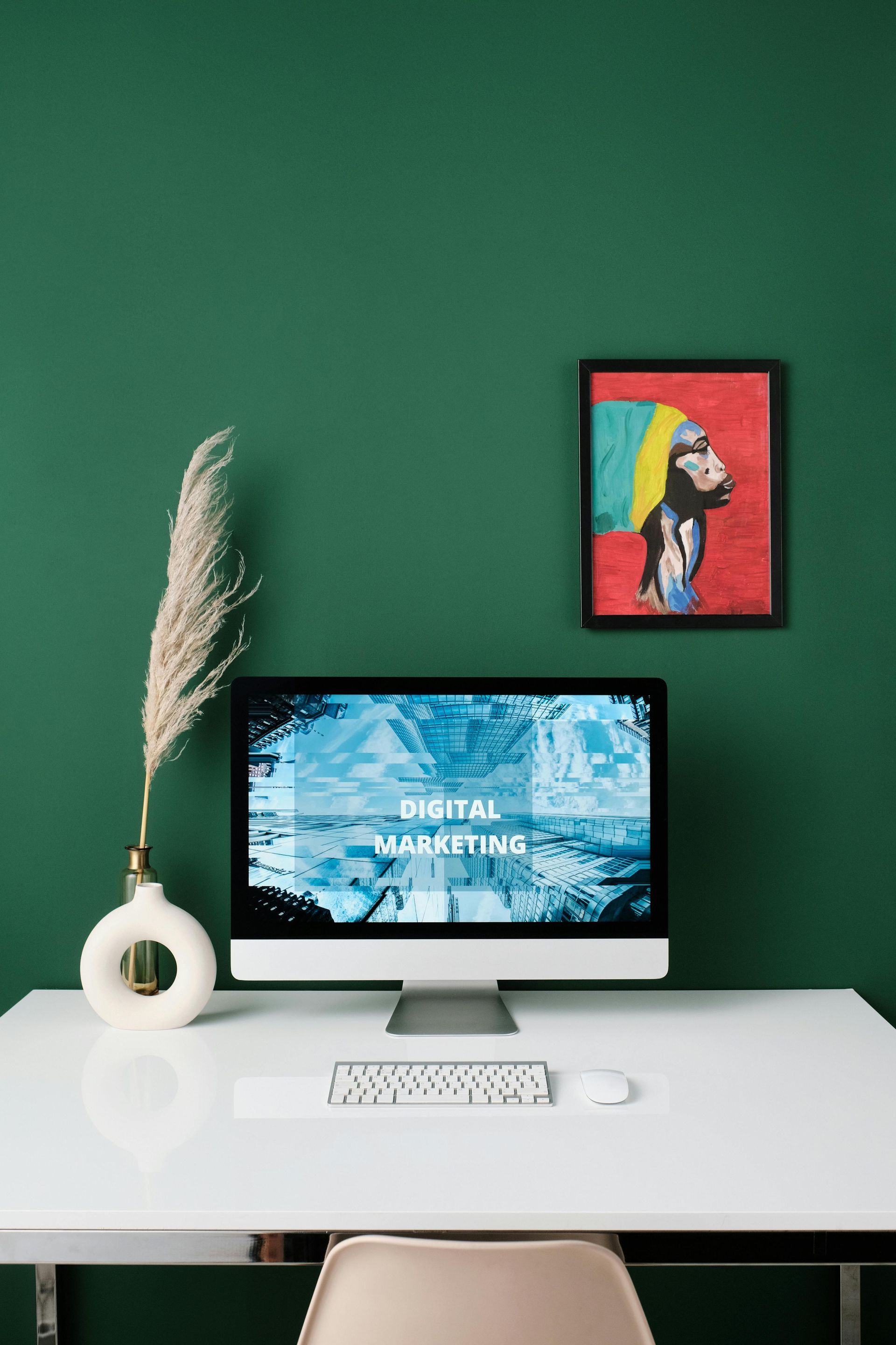 Desktop computer displaying "Digital Marketing" text, on a white desk against a green wall; vase with plant, artwork.