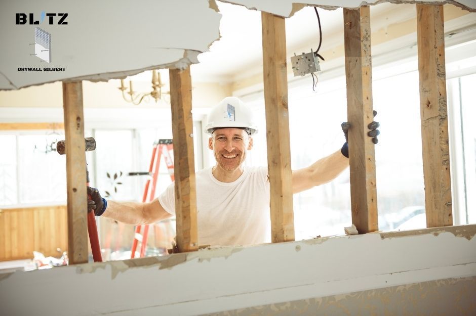 A drywall repair company professional in a white hard hat and t-shirt smiles while working on a residential renovation project. He wears black gloves and stands among exposed wooden framing with damaged drywall and electrical fixtures visible near W Golden Ln, and N 54th Ave in Glendale, AZ.