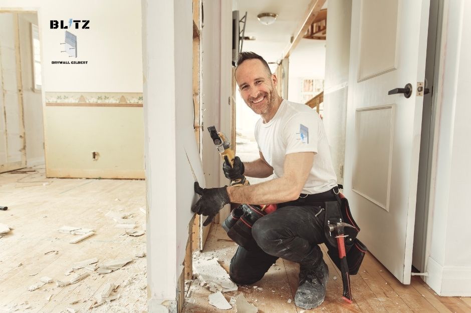 A drywall repair company worker wearing white t-shirt and dark pants, kneeling while holding a recip saw. The technician is smiling brightly in a residential construction site with white walls and appears to be working on wall repair near W Green Hollow Terrace, and N Clear Creek Dr, Litchfield Park, AZ.