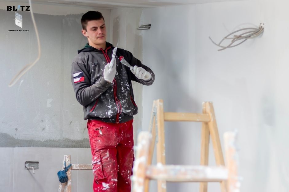 A drywall repair company worker wearing red pants and a gray hoodie is repairing a residential indoor wall. He holds a hand tool, likely a scraper or putty knife, and is smoothing plaster with white dust on his clothes and gloves near E Maddock Rd, and N 7th St, New River, AZ.
