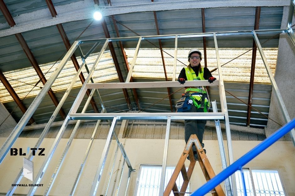 A worker for a drywall repair company in a safety gear is working with a metal framing installation in a room with exposed wooden studs and bright lighting. This drywall contractor is servicing a commercial job site near S Western Ave, and W Ray St, Superior, AZ.