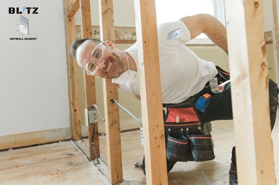 A drywall repair contractor wearing safety glasses and a white shirt works on a residential construction project while kneeling among wooden framing. He wears a tool belt and is working near W Monterey Way, and N 126th Dr in Avondale, AZ.