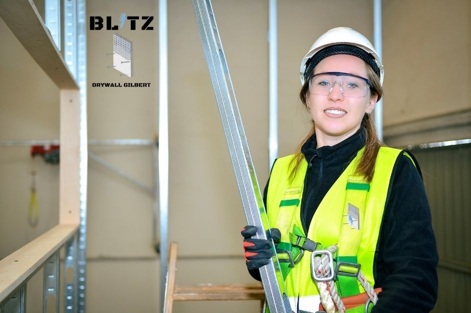 A drywall repair contractor wearing a white hard hat and high-visibility yellow safety vest stands in a commercial building interior, holding a metal framing for drywall installation project near W Press Rd, and N Hearn St in Queen Creek, AZ.