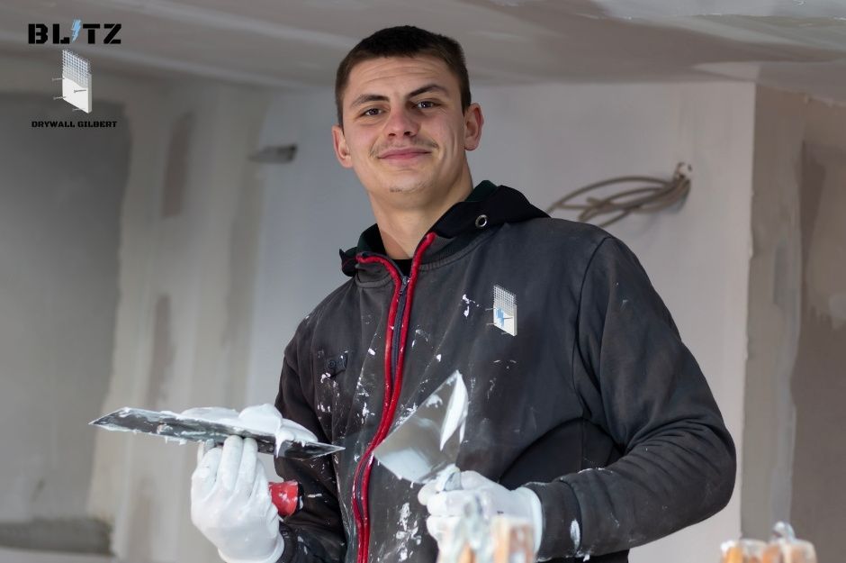 A drywall repair contractor smiles while holding a putty knife and wearing white gloves, with white compound splattered on his dark jacket. He stands in an unfinished room with white walls during a residential project near N 43rd Ave, and W Twin Peaks Ln in Rio Vista, AZ.