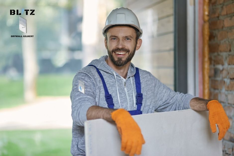 A drywall repair contractor in white hard hat smiles while holding white drywall sheets on a residential construction site. He is wearing orange work gloves. The background shows a home under renovation with natural lighting near N 112th Ln, and W Nevada Ave in Youngtown, AZ.