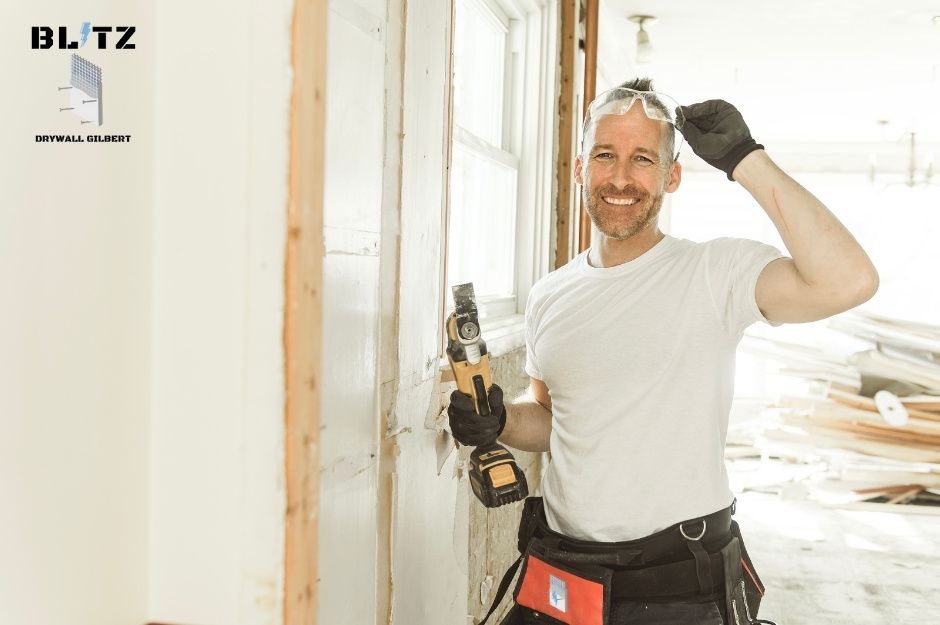 A drywall repair handyman in a white t-shirt and work gloves holds a reciprocating saw while adjusting his safety glasses in a commercial renovation space. He wears a tool belt and stands near windows with exposed framing and construction materials near S 25th Ave, and W Jessica Ln in Liberty, AZ.