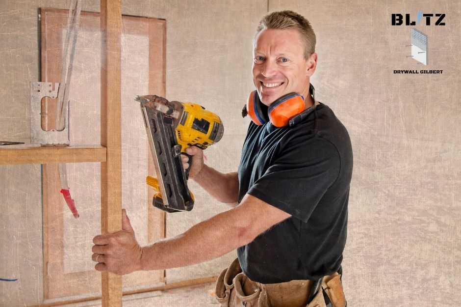 A smiling drywall repair specialist wearing an orange ear protection and a black shirt holding a yellow nail gun is working in a commercial construction site with wooden framing and drywall visible behind him near N Motoya Ln, and W Descanso Canyon Dr, Casa Grande, AZ.