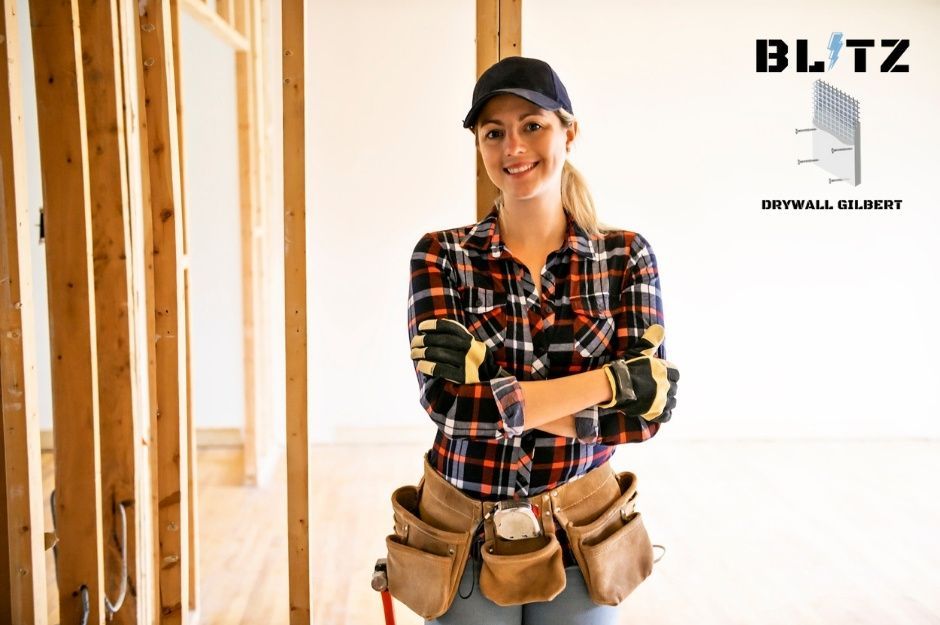 A smiling drywall repair specialist woman wearing a black baseball cap and red plaid shirt, with a tool belt around her waist containing various hand tools, stands confidently with arms crossed in a commercial construction area with wooden framing visible behind her near W Merriweather Wy, and N Hudson Dr, Florence, AZ.