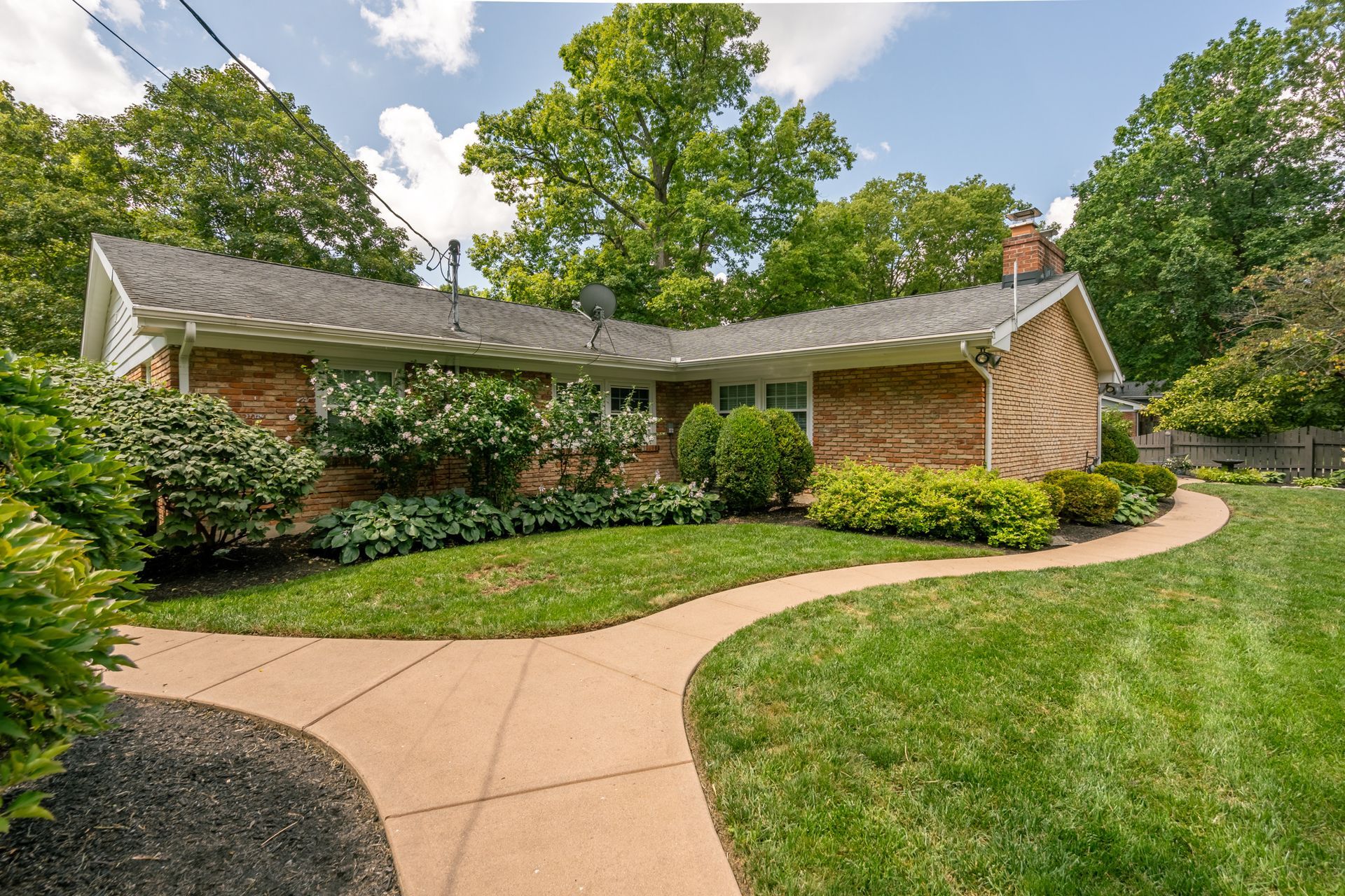 Brick ranch-style house with curved walkway through green lawn, surrounded by trees.