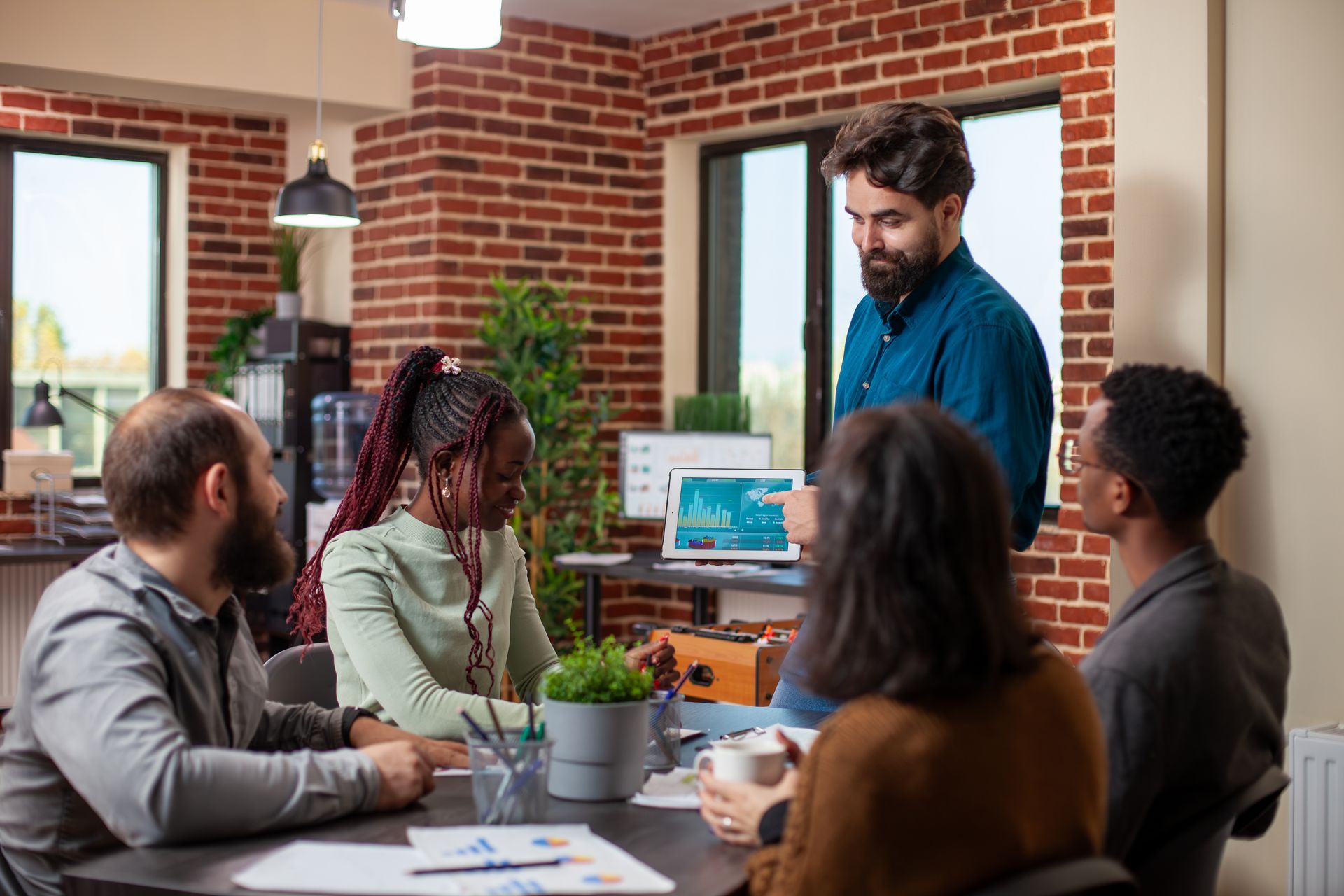 Business team in a meeting around a table, one person showing a tablet. Red brick wall.