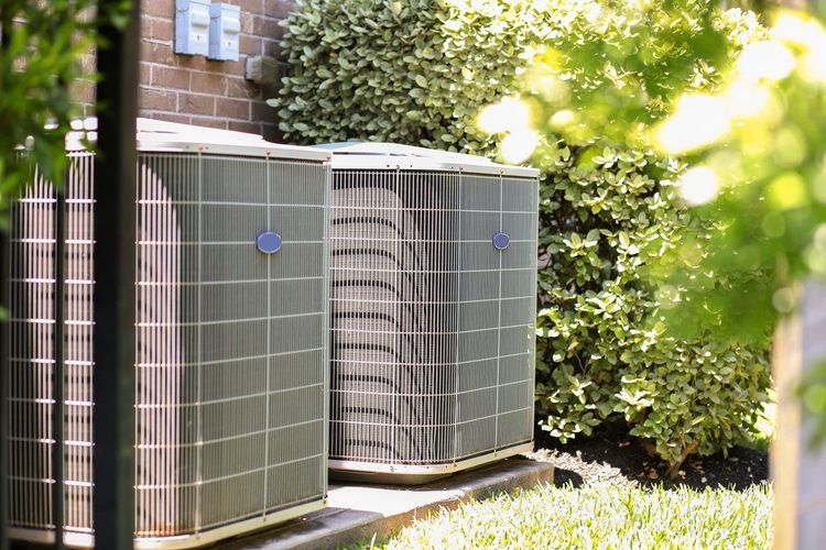 Two outdoor air conditioning units next to a brick building and surrounded by greenery.