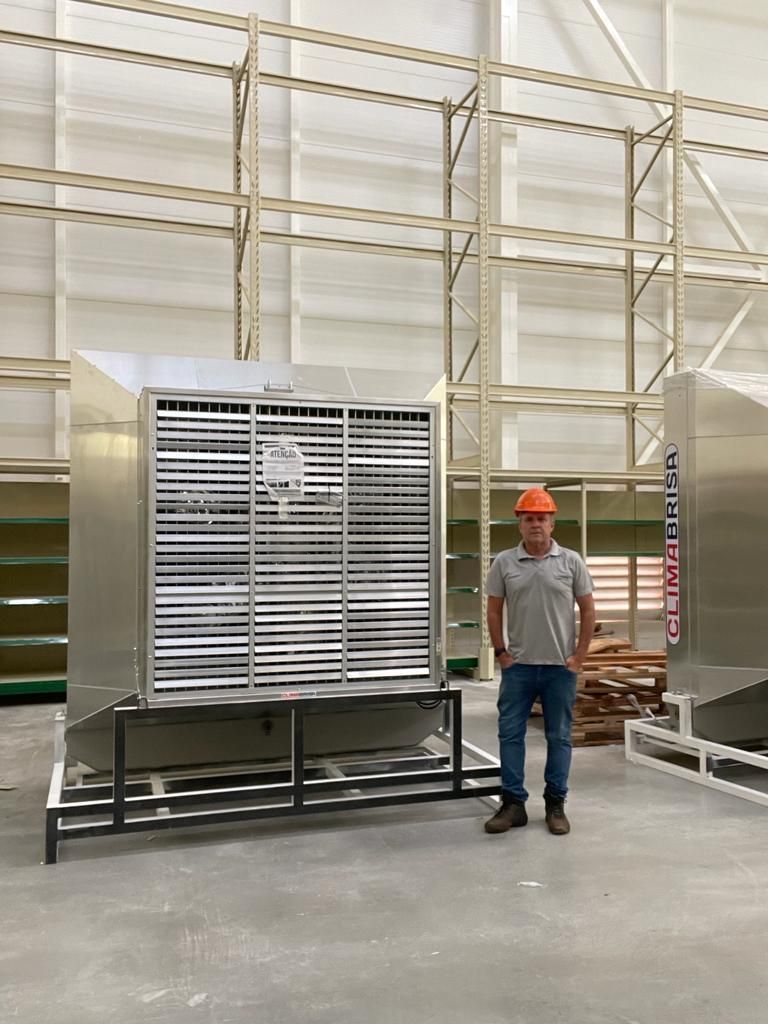 Uma pessoa com capacete de segurança está ao lado de um grande ventilador industrial em um armazém com estantes metálicas.