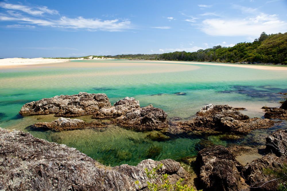 A Body Of Water With Rocks In The Foreground And A Beach — Holiday Coast Pools & Water In Sawtell, NSW