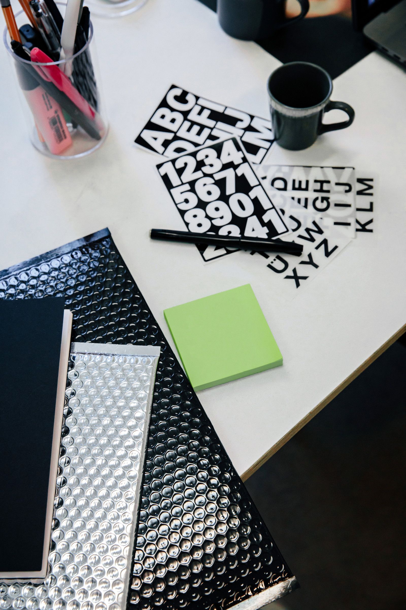 Workspace with alphabet stencils, a green notepad, a black mug, and stacked patterned folders on a white desk