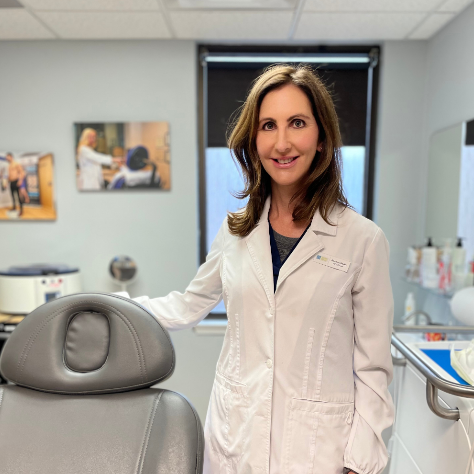 Woman in a white lab coat standing beside a dental chair in a bright clinic.