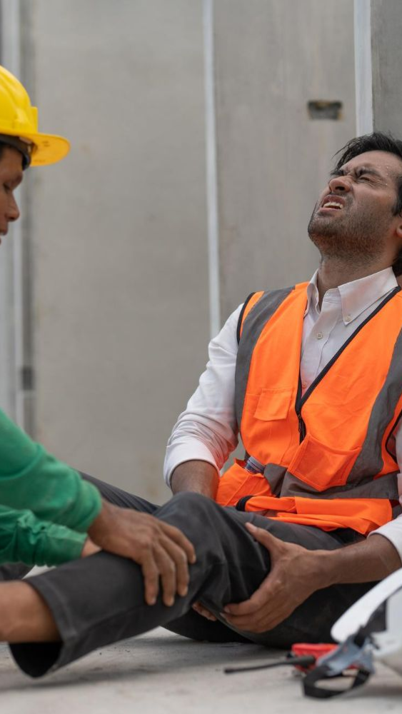Construction worker injured on the floor, being helped by another. They are in a building.