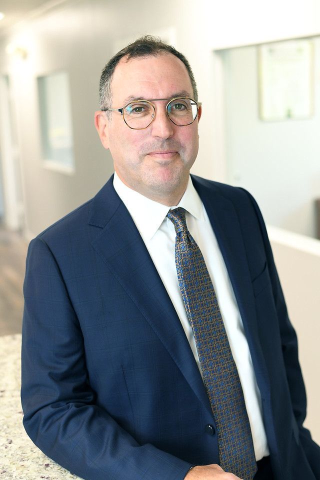 Man in suit and glasses, leaning against a counter, smiling in an office setting.