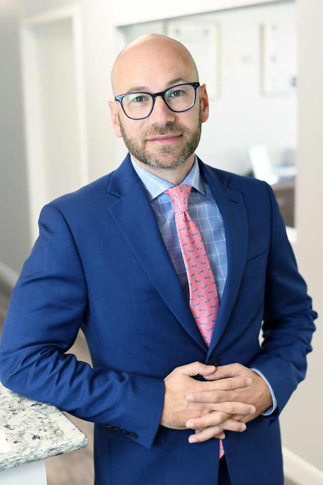 Man in blue suit, glasses, and pink tie, arms crossed, leaning on counter.