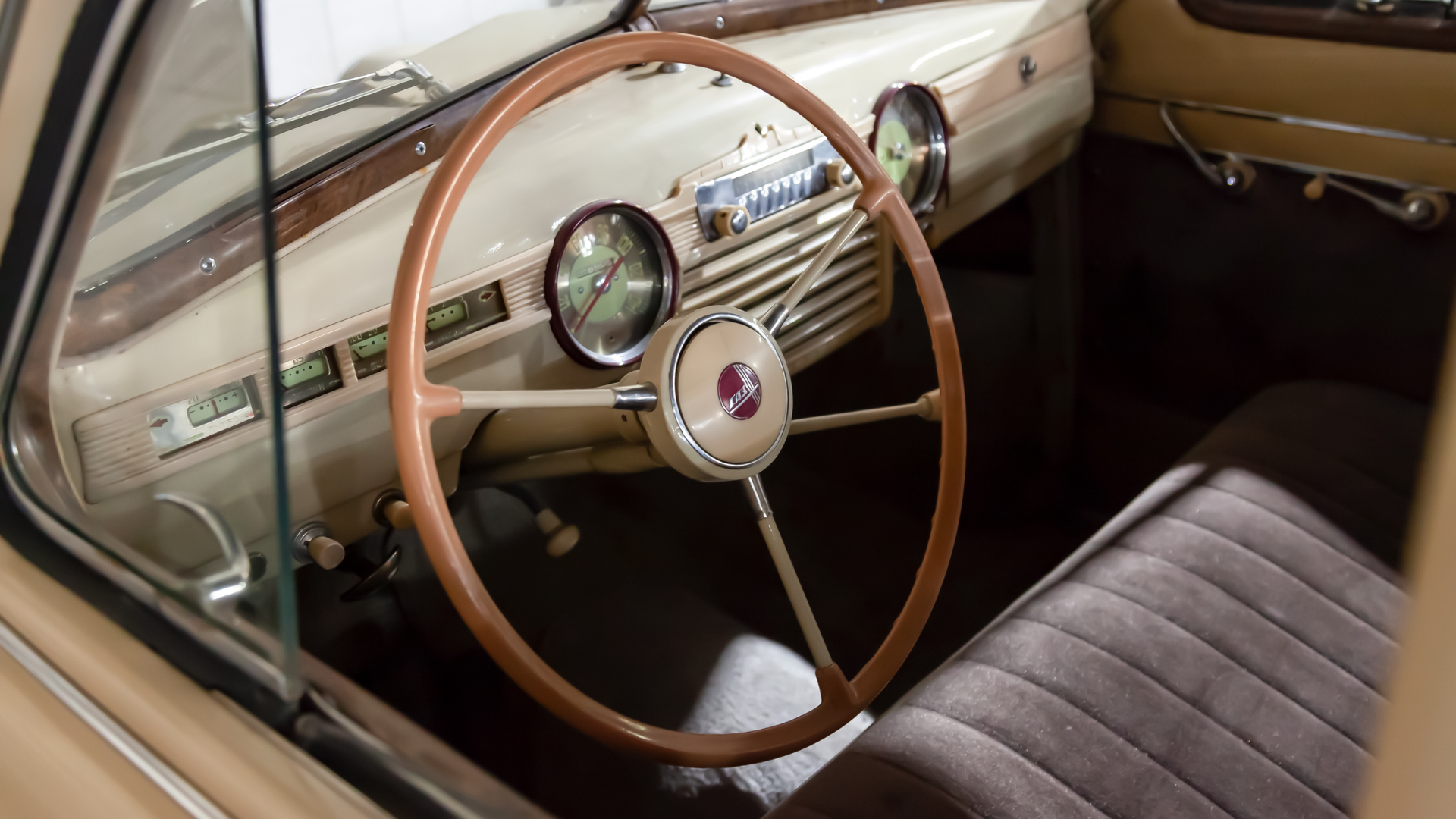Interior of a classic beige car with brown steering wheel, dashboard, and gauges. Dark seat.