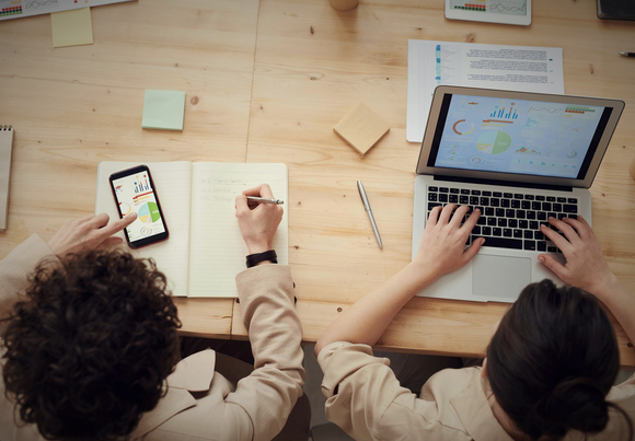 Deux personnes collaborent à un bureau en bois, utilisant un ordinateur portable et un smartphone, avec des notes et un crayon à portée de main.