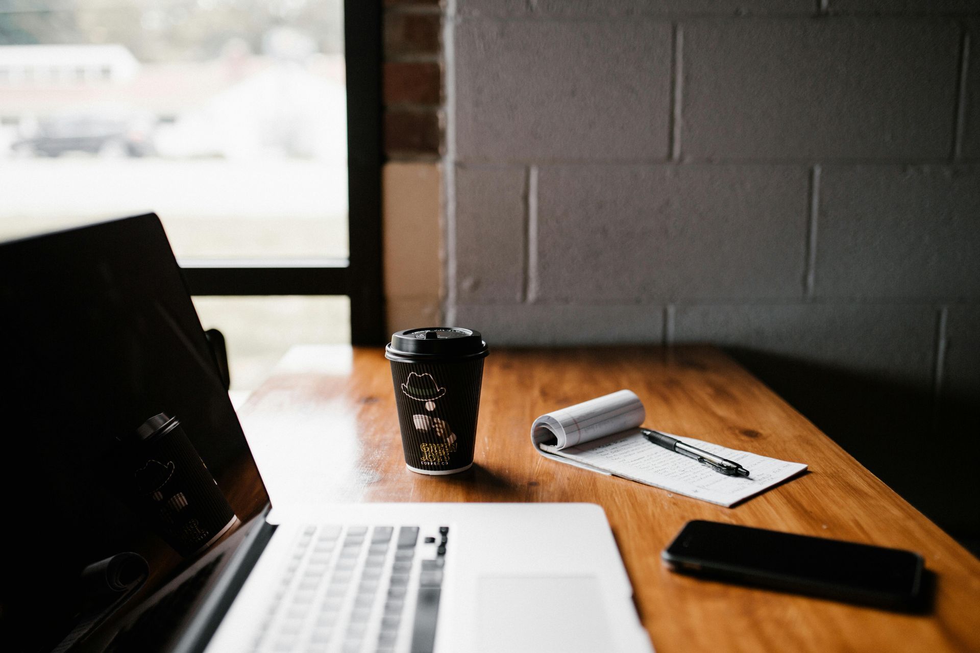 Un ordinateur portable posé sur un bureau en bois, à côté d'une tasse de café, d'un carnet et d'un téléphone près d'une fenêtre.
