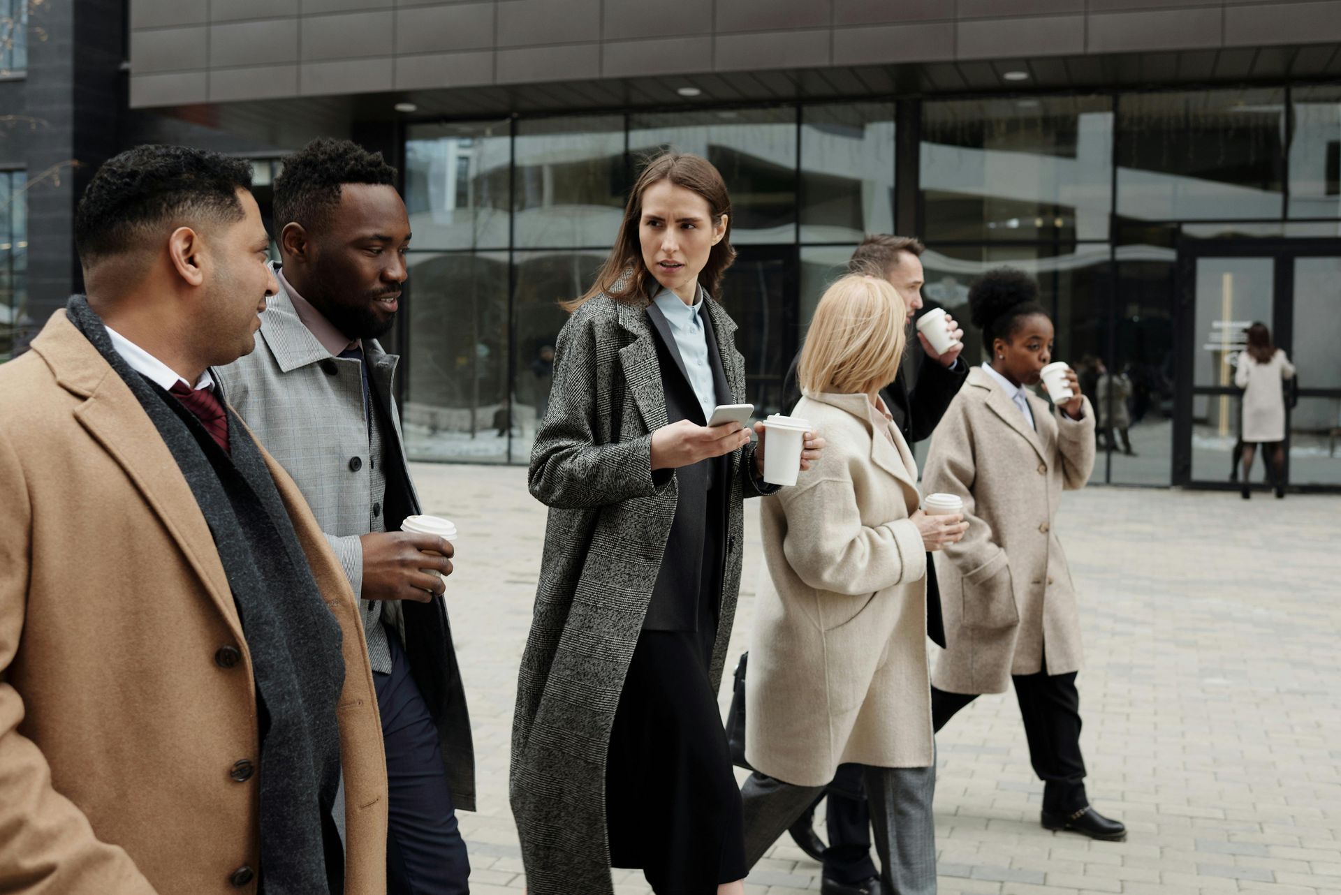 Des personnes marchent devant un immeuble de bureaux moderne en verre, plusieurs tenant des tasses de café.
