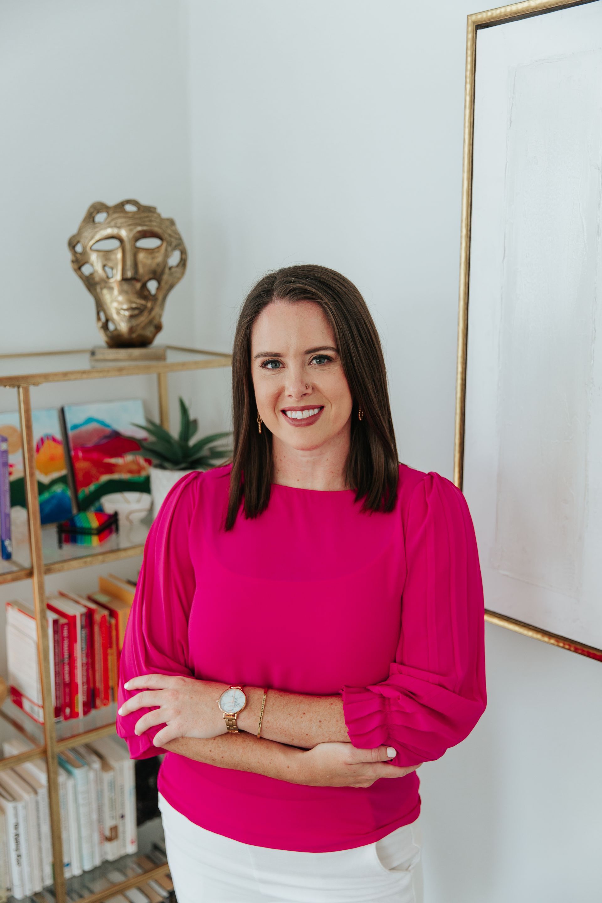 A woman in a pink sweater and white skirt is standing in front of a bookshelf.