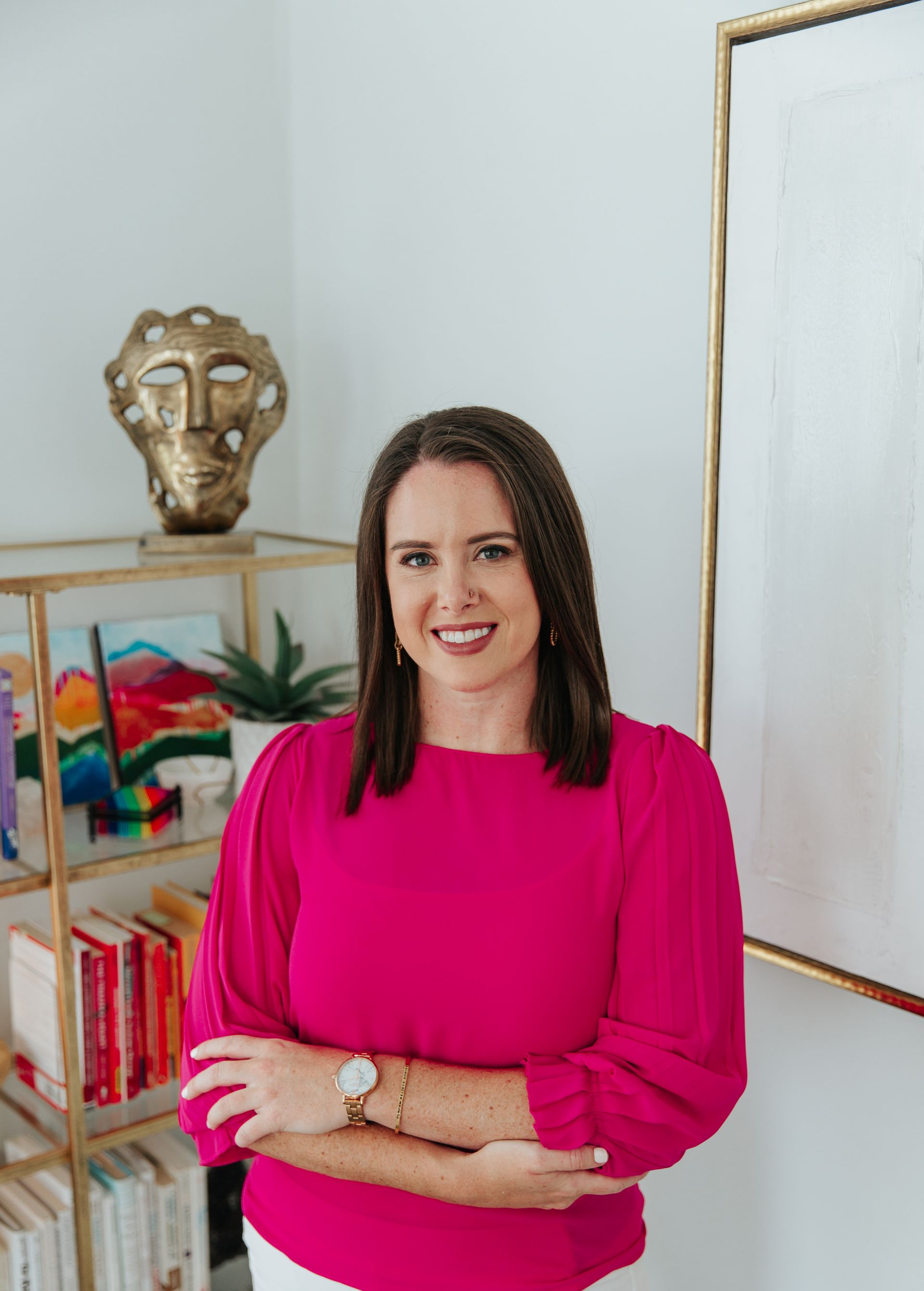 A woman in a pink sweater and white skirt is standing in front of a bookshelf.