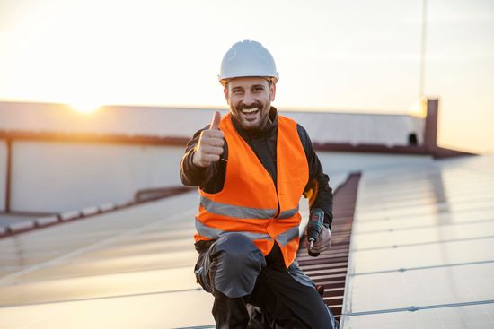 Worker in orange safety vest and white hard hat giving a thumbs-up on a rooftop at sunset.