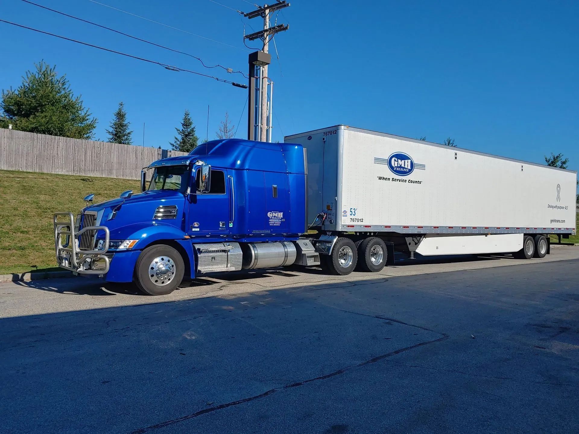 Blue semi-truck with a white trailer parked on a paved surface in a sunny setting.