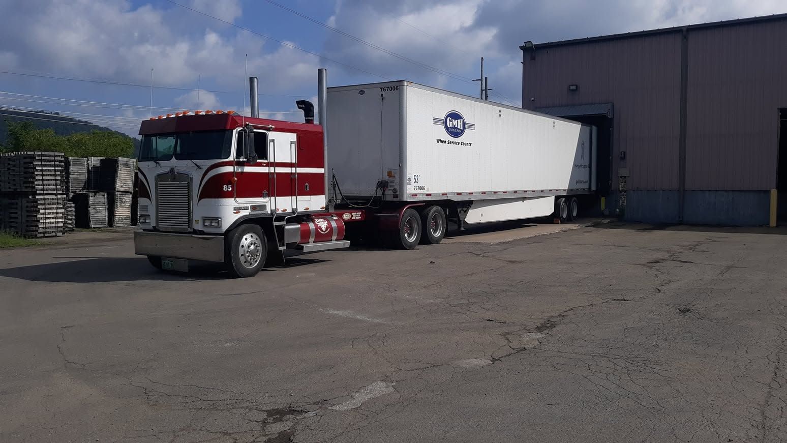 Red semi-truck with white trailer parked beside a warehouse loading dock.