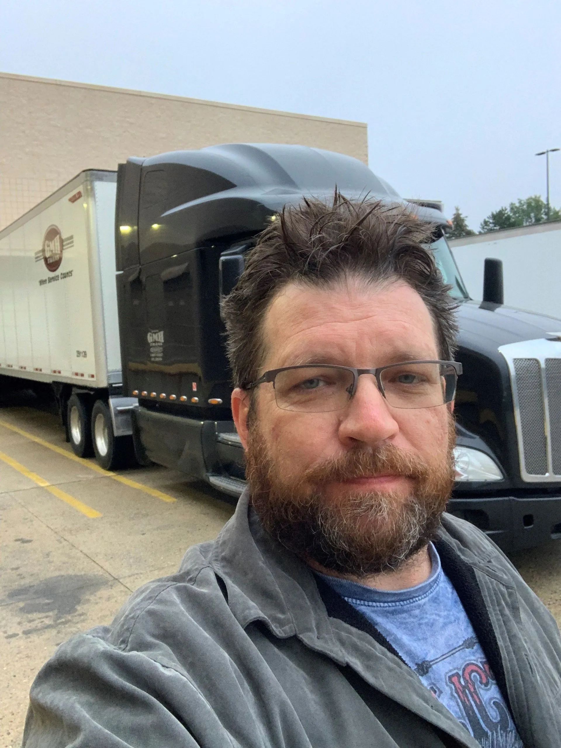 A man with a beard is taking a selfie in front of a semi truck.