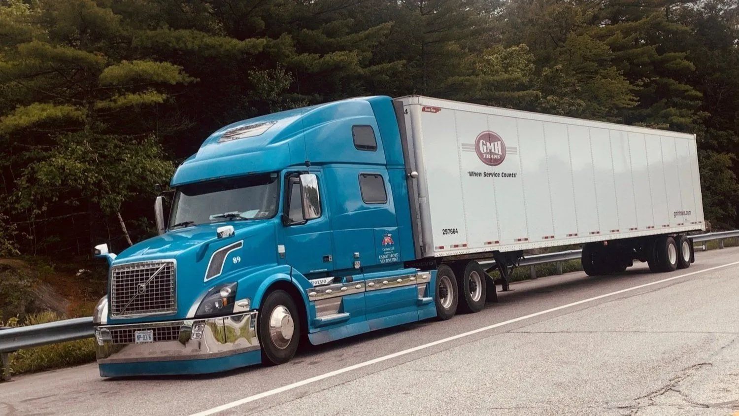 Blue semi-truck with white trailer on a road. Trees in the background.