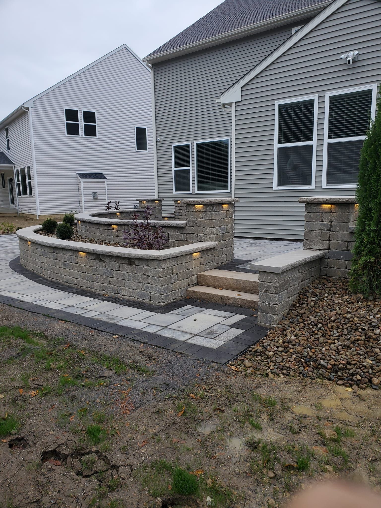 Stone patio with retaining walls, built-in fire pit, steps, and up-lighting, next to two houses.