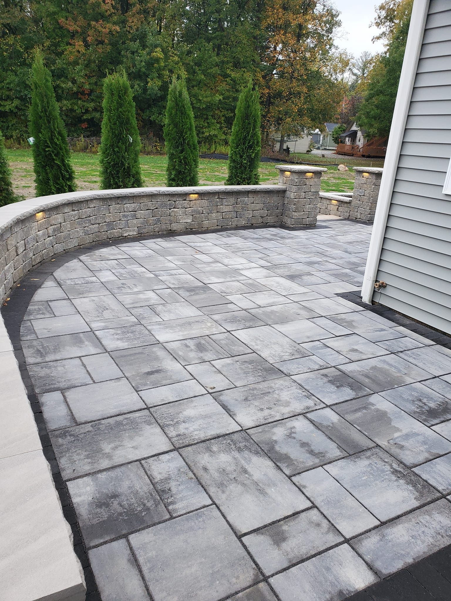 Patio paved with grey blocks, bordered by a stone wall. Green trees in the background, near a house.