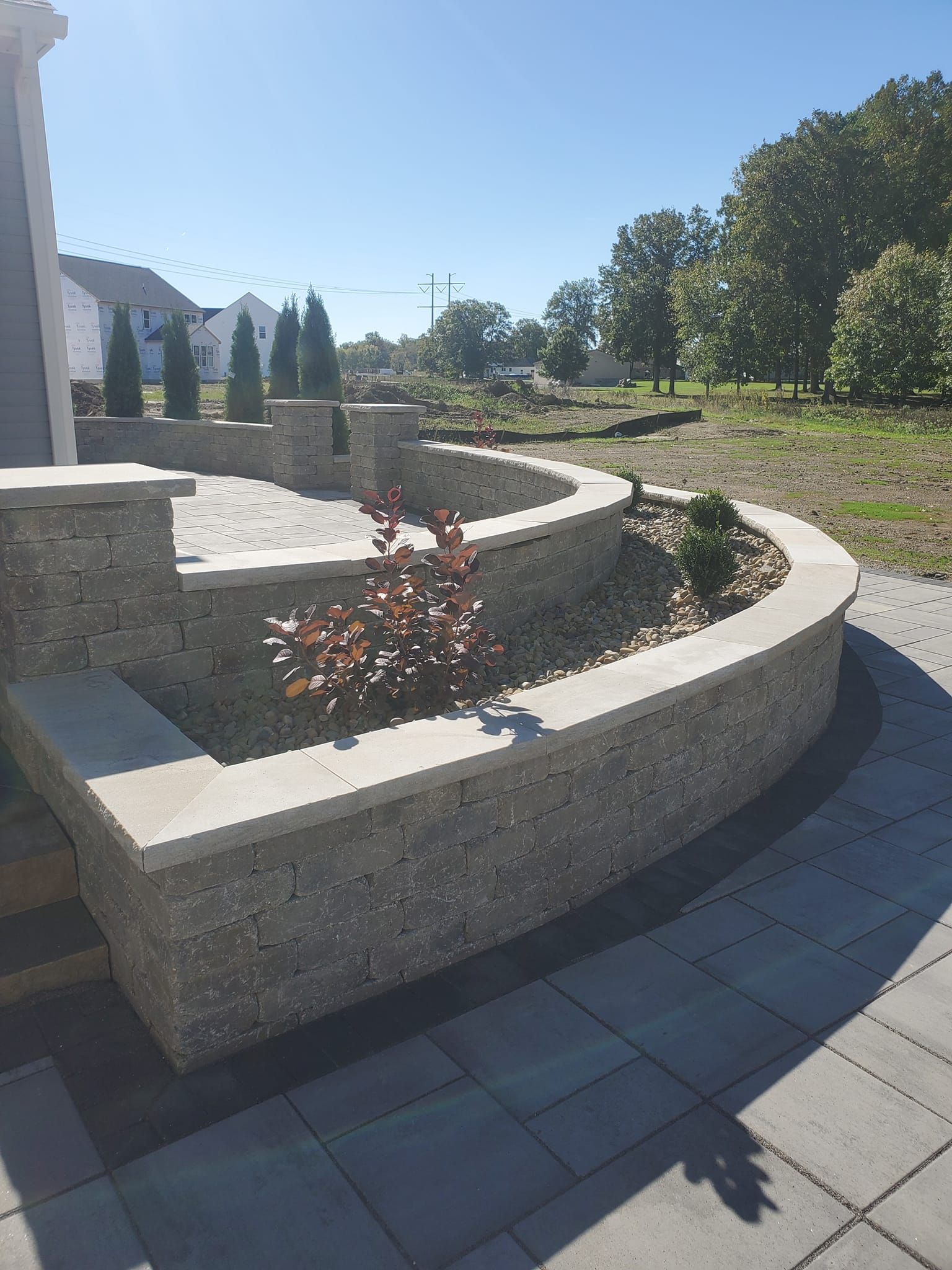 Curved stone retaining wall with a patio, steps, and landscaping under a sunny sky.