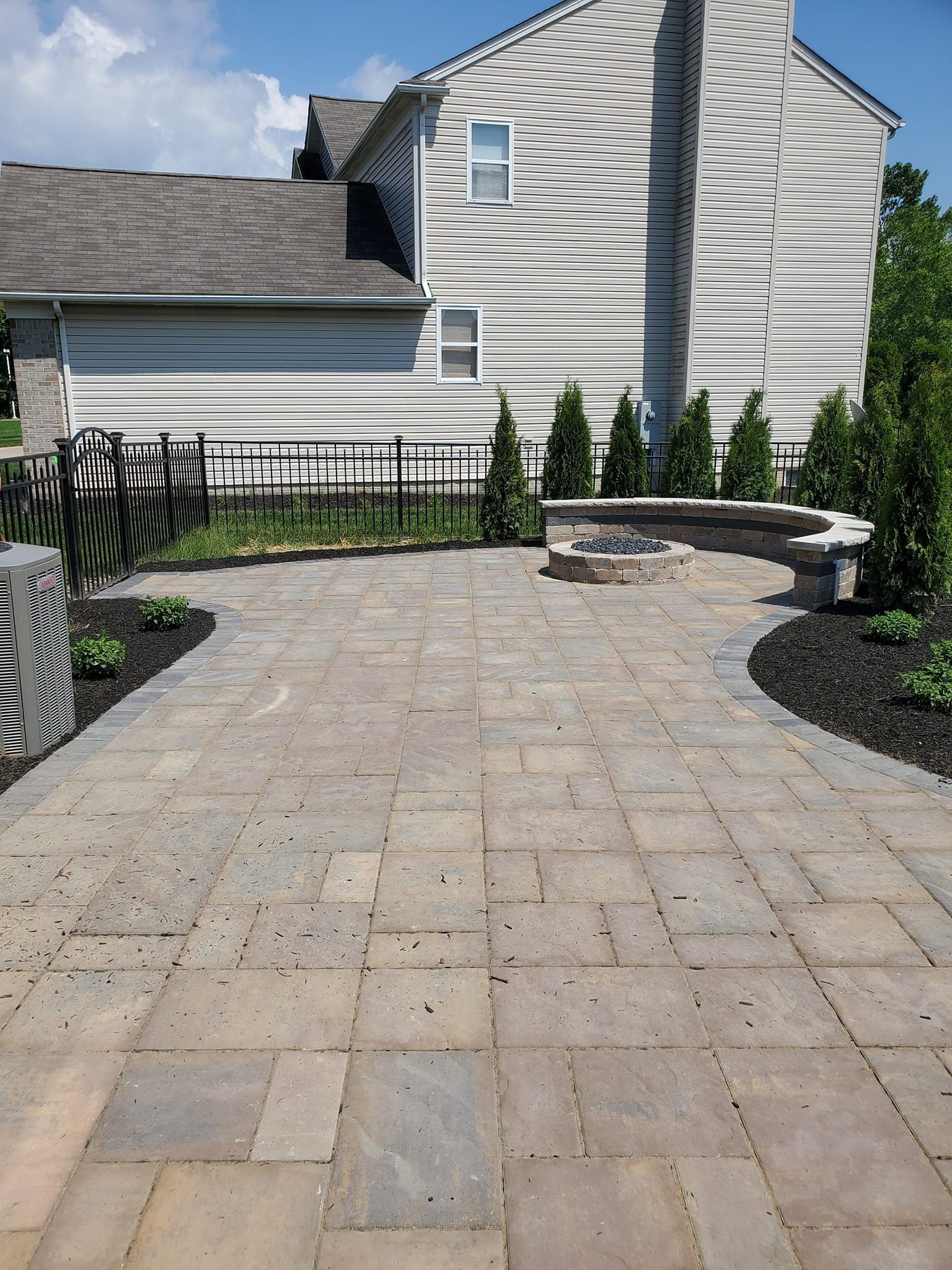 Paver patio with a fire pit, adjacent to a two-story house with green landscaping.