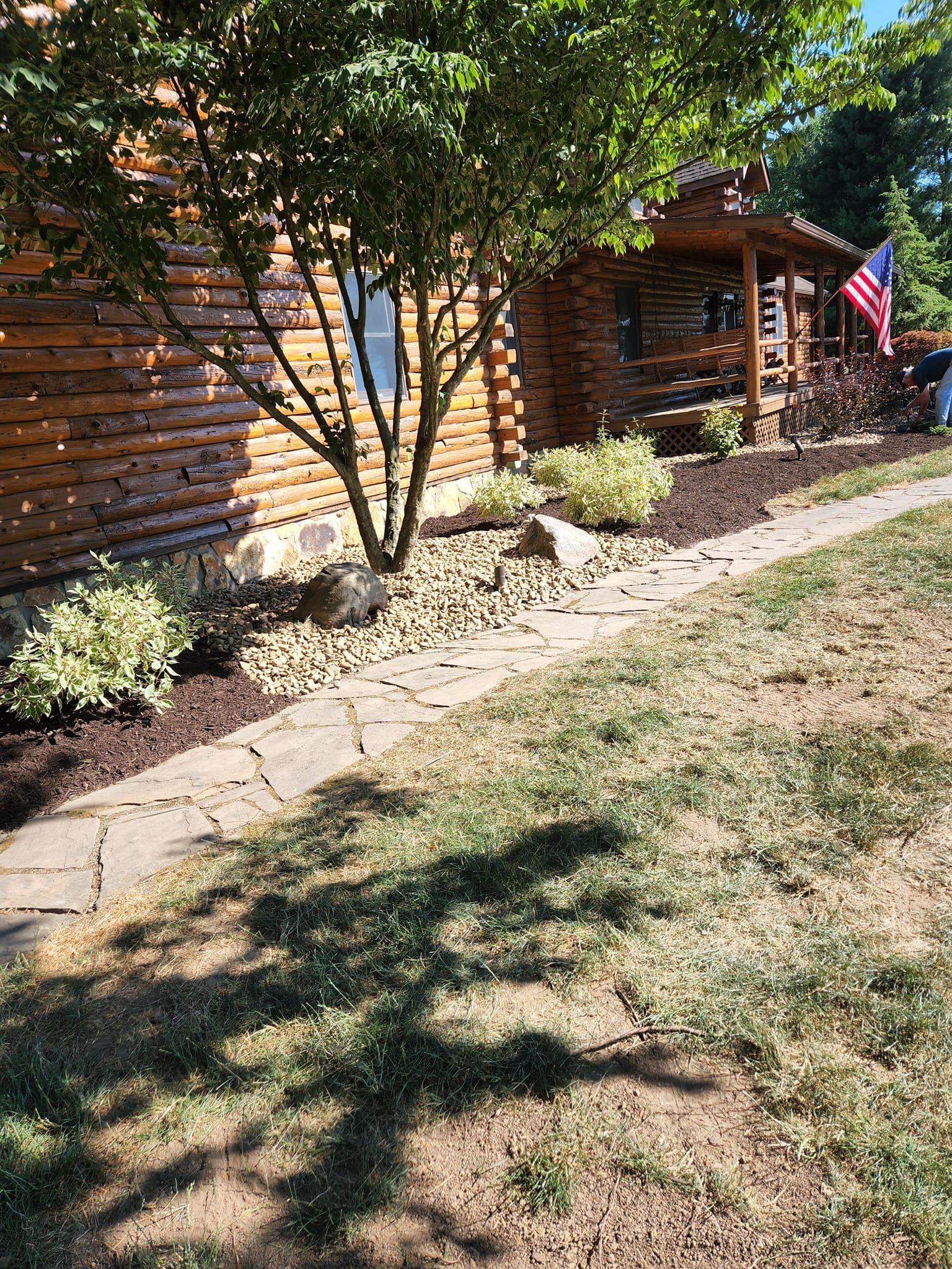 Log cabin with flag, porch, and a stone path along a landscaped bed with a small tree.