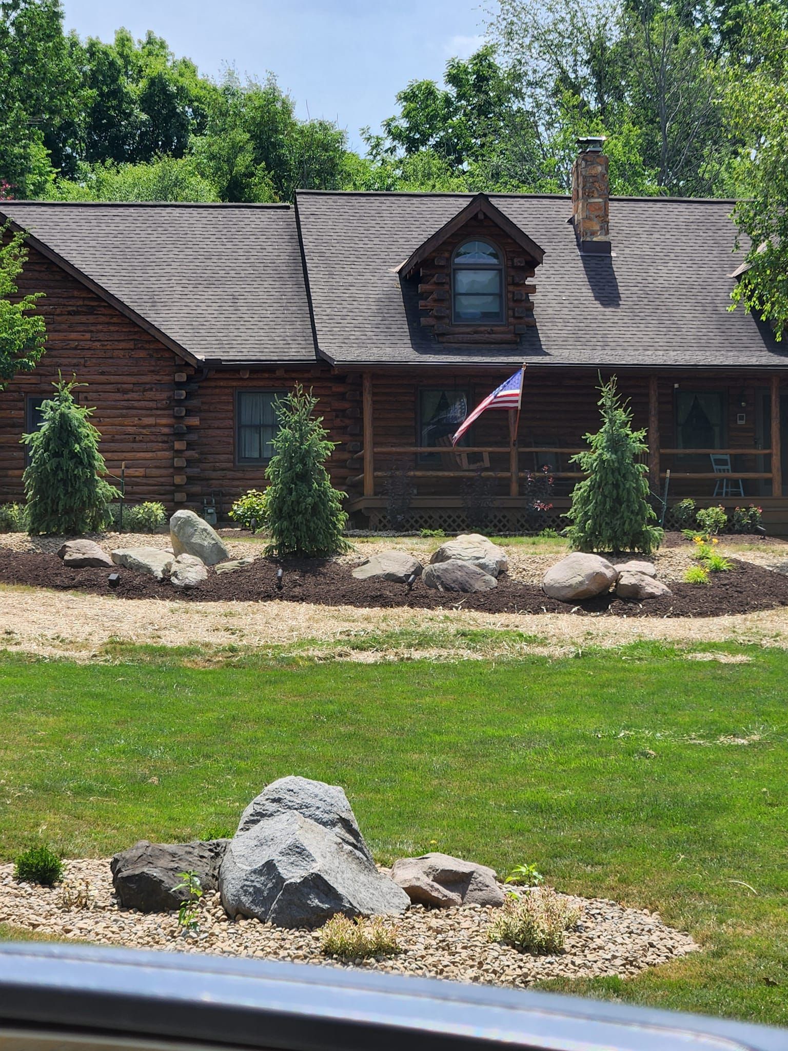 Log cabin with American flag, trees, and landscaping in front. Green grass and blue sky.
