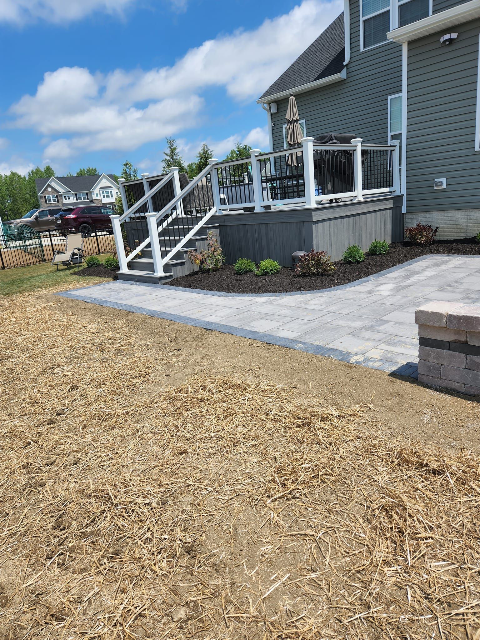 Backyard deck with stairs and paver patio, next to a house under a blue sky.