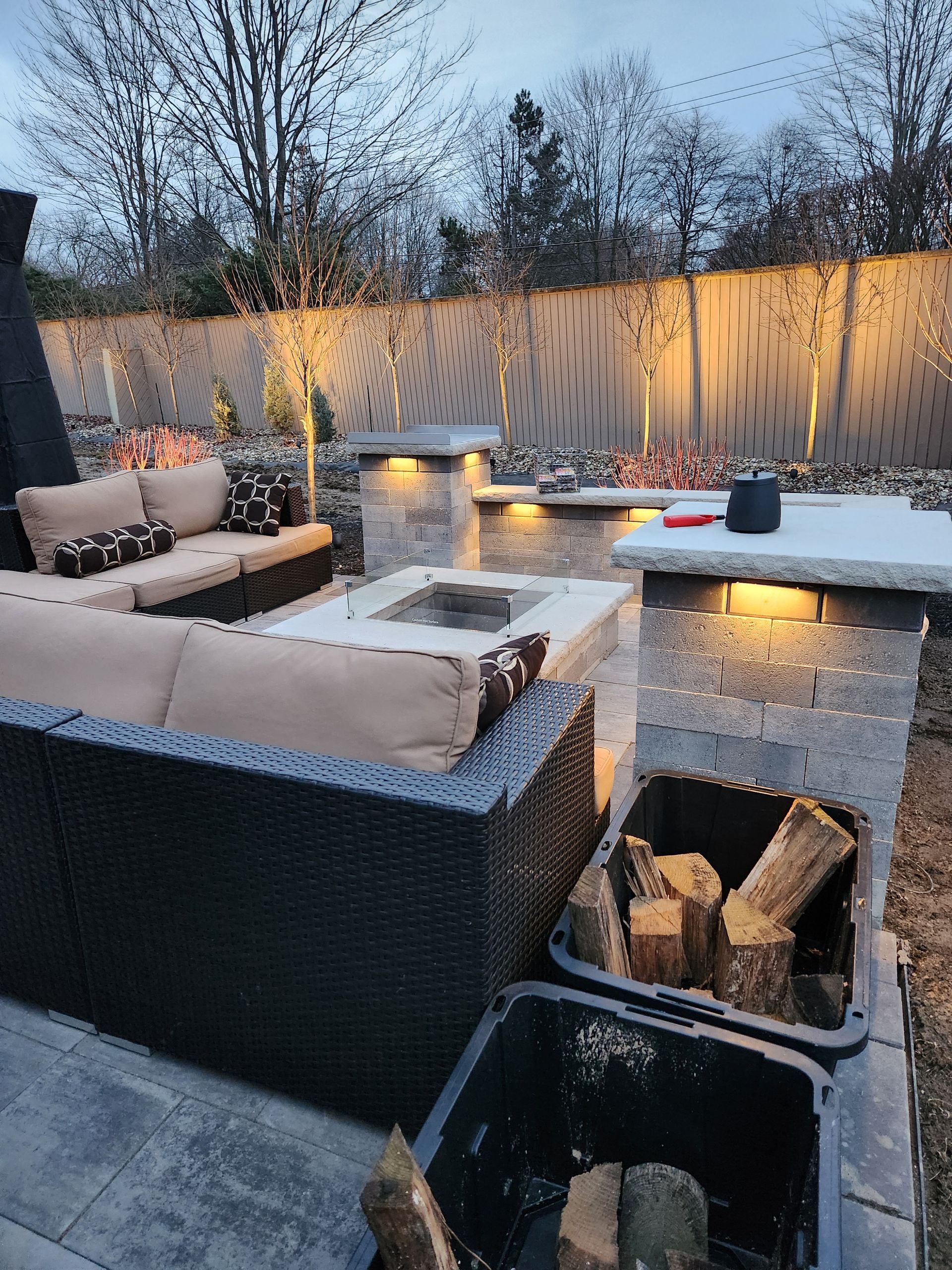 Outdoor patio with wicker furniture, fire pit, and stonework, lit by ambient lighting. Firewood in a bin.